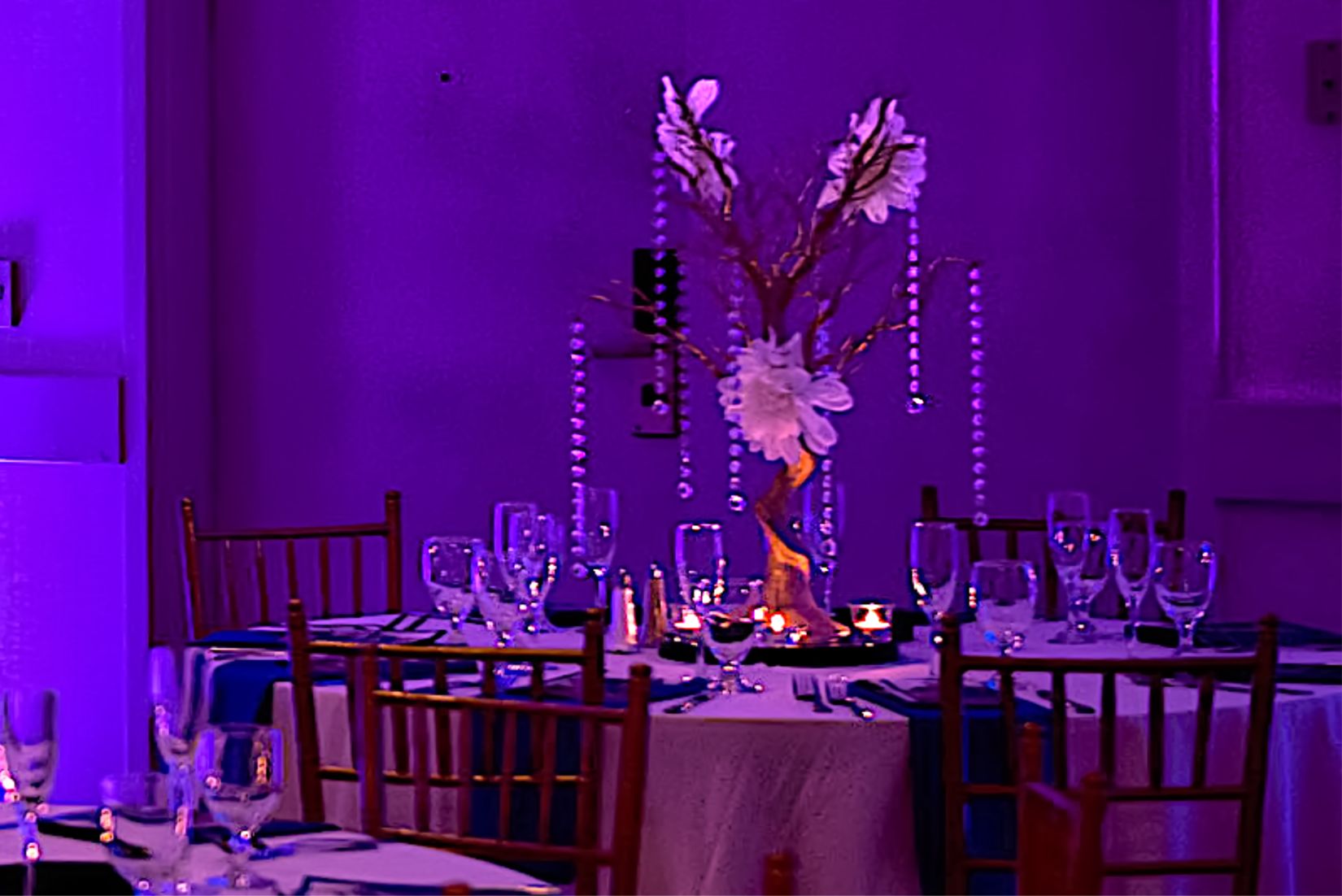 Beautiful gold manzanita tree centerpiece accented with white flowers and hanging crystals. Event decor features purple uplighting and chiavari chairs.